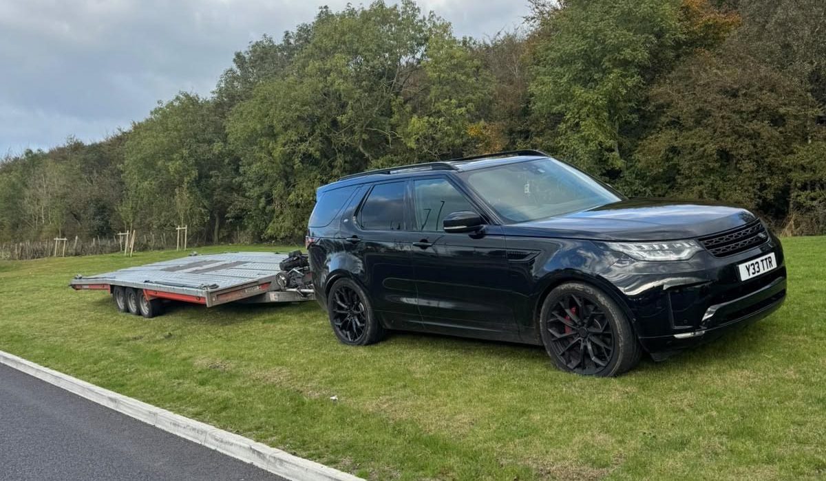 The TTR Recoverys black Land Rover Discovery hitched to its tri-axle car transporter trailer, parked on grass by a road